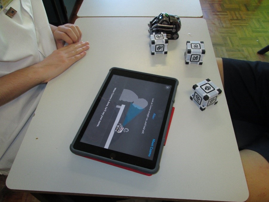 Image of the robot in action. There is an iPad set up with the Cozmo app. On a table there are three cubes that have been activated, as per the previous instructions, and there is the Cozmo robot.