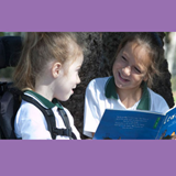 Image of two girls reading a book, one is sitting in a motorised scooter